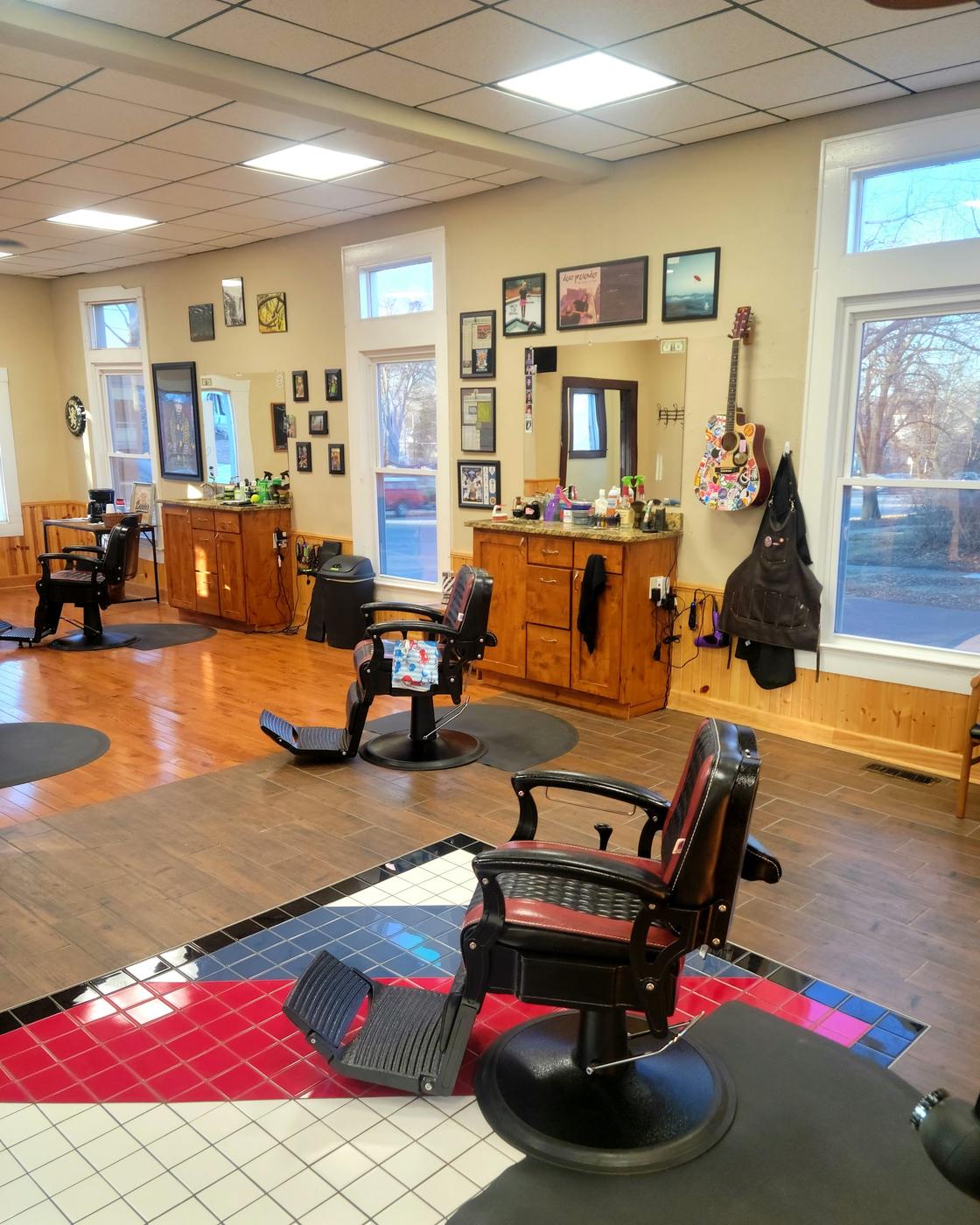 The cutting floor: two red and black barber chairs, wooden stations with granite tops and mirrors, a sticker covered guitar on the wall, and a red, white and blue tiled floor.