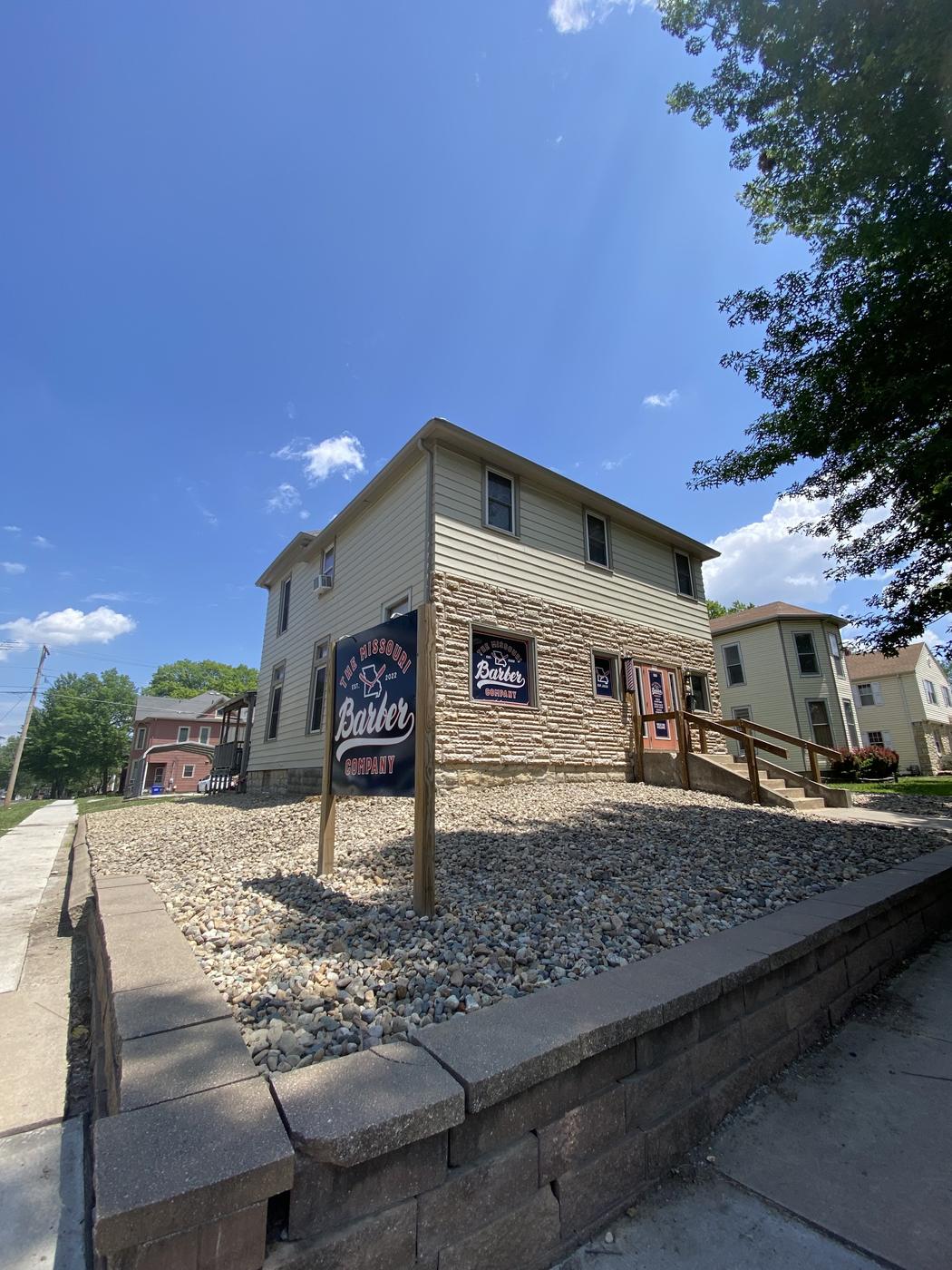 The Missouri Barber Company on the corner of West Broadway Boulevard in Sedalia: a two story building in cream siding over limestone, with a navy yard sign and a barber pole by the door.